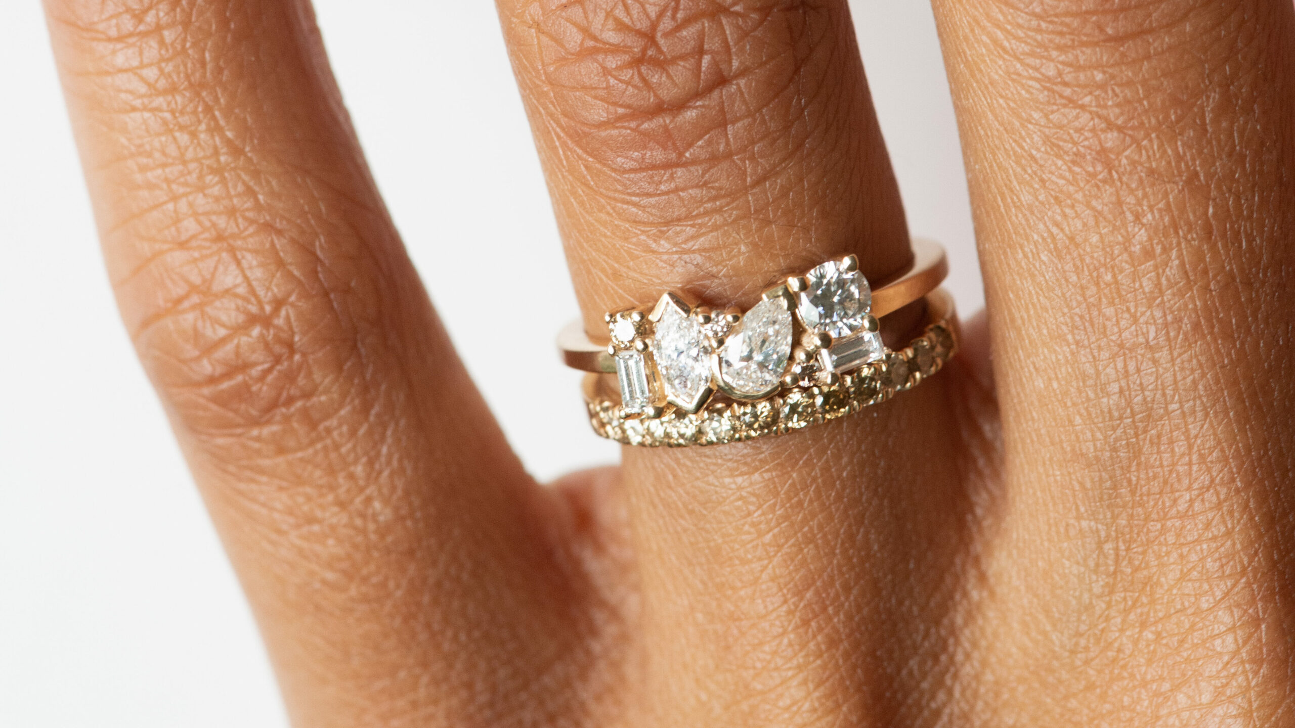 Close up of a hand wearing two rings; One linear cluster diamond ring, where each diamond is set in a different cut, and another linear yellow sapphire band.