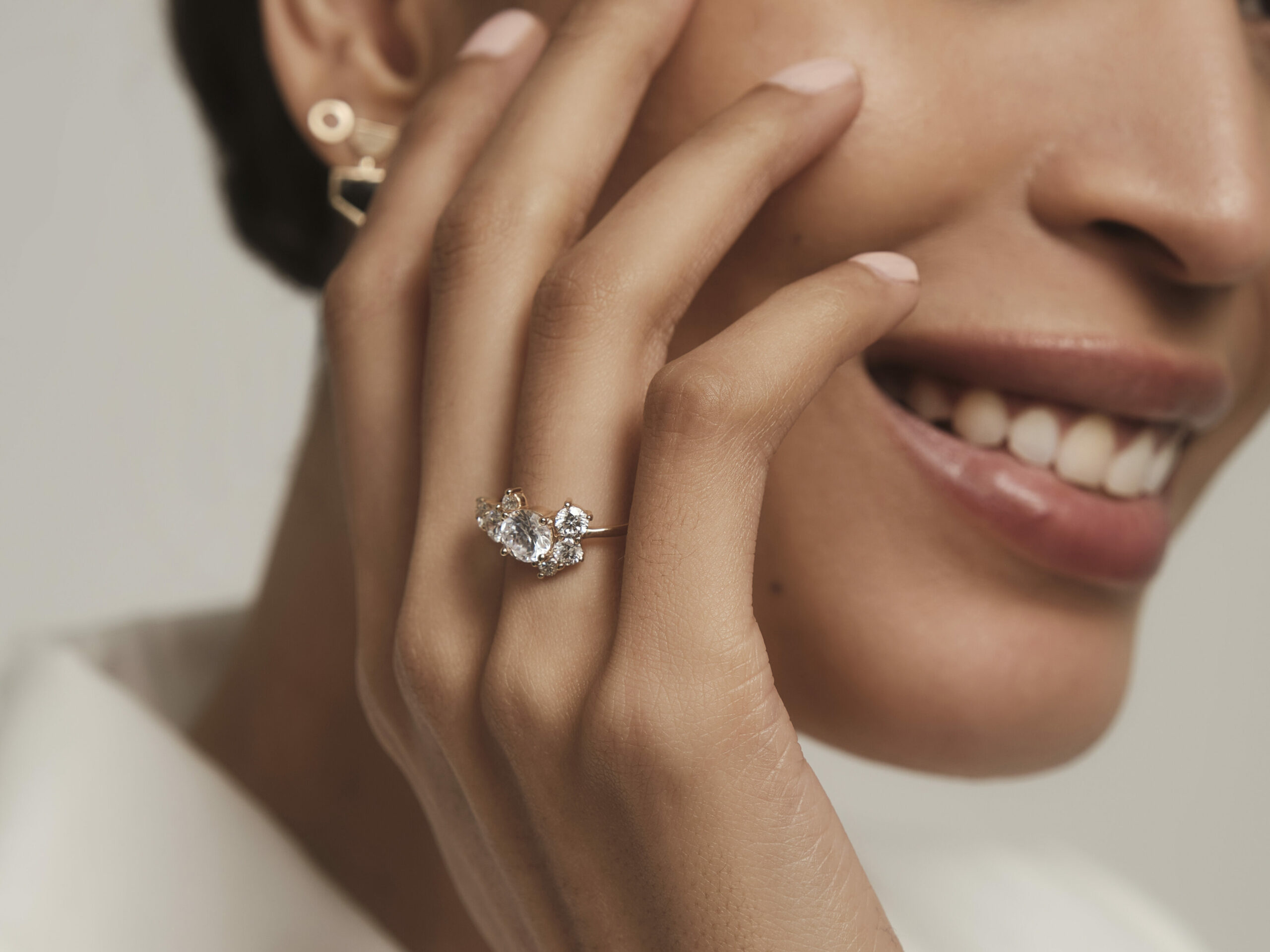 Hand placed gently on model's cheek. Ring finger wearing a Burst Cluster Diamond Ring. Model smiling, hand in focus