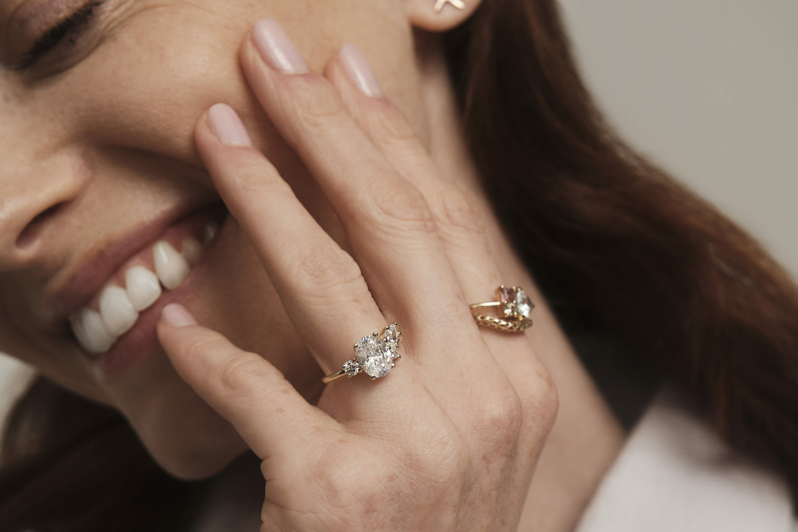Hand placed gently on model's cheek. Pointer finger wearing Eaves Cluster ring, ring finger wearing a filagree and Charta cluster stack. Model smiling, hand in focus