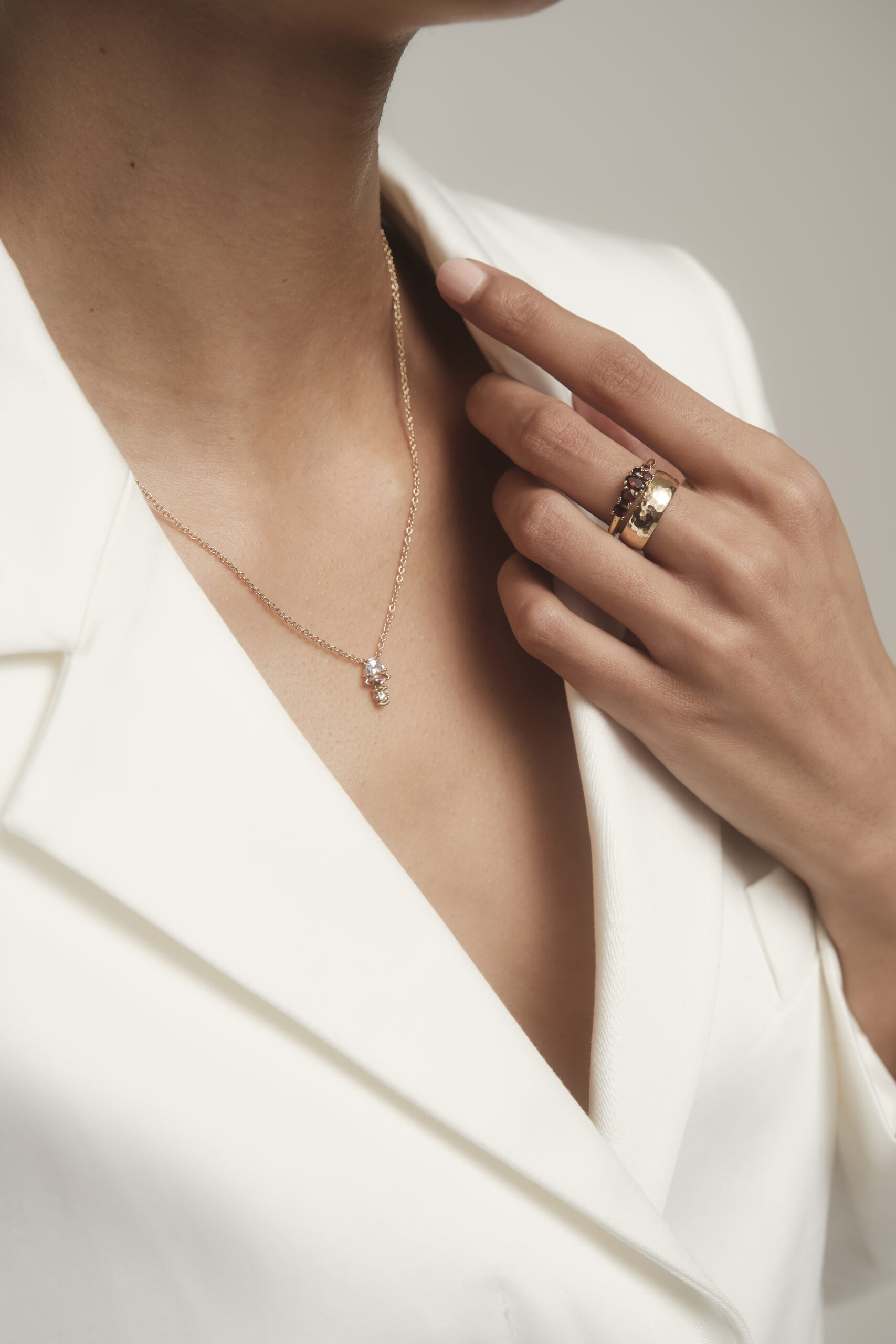 woman gracefully placing hand on blazer collar, showcasing stack of rings on middle finger. She is also wearing a gemstone and diamond pendant necklace.